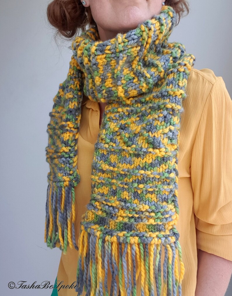 May include: A knitted scarf with a yellow, green, and gray color pattern. The scarf has long fringe tassels on both ends.