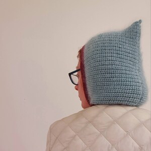 Pu&ograve; includere: Un cappello all'uncinetto azzurro con la parte superiore a punta. Il cappello ha un aspetto strutturato e viene indossato sopra una testa con gli occhiali. La persona indossa una giacca trapuntata bianca. L'immagine include il testo "Tasha BoSpoke".