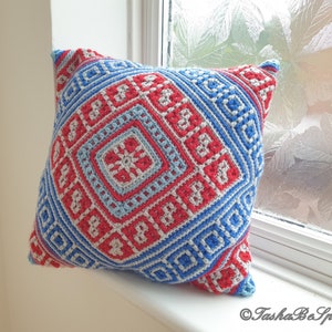 Op de afbeelding: Een vierkant gehaakt kussen met een geometrisch patroon in rood, blauw en lichtblauw. Het kussen heeft een centraal vierkant ontwerp met een bloemachtig motief, omgeven door herhalende ruit- en vierkantvormen. Het kussen ligt op een witte vensterbank.