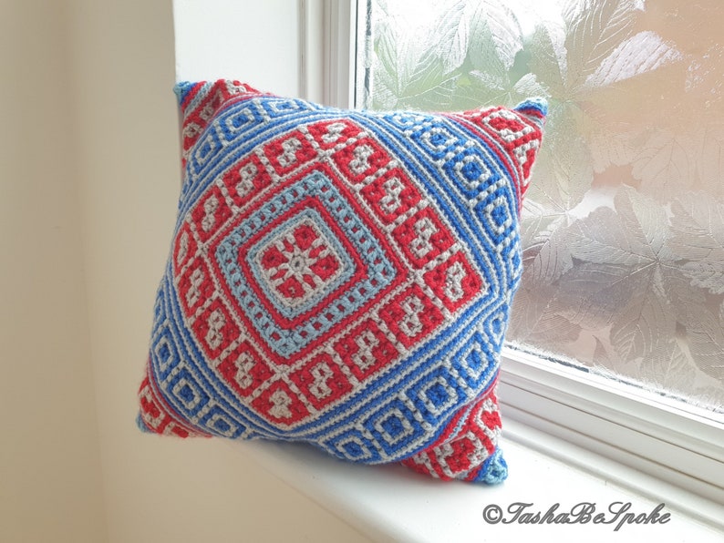 Op de afbeelding: Een vierkant gehaakt kussen met een geometrisch patroon in rood, blauw en lichtblauw. Het kussen heeft een centraal vierkant ontwerp met een bloemachtig motief, omgeven door herhalende ruit- en vierkantvormen. Het kussen ligt op een witte vensterbank.