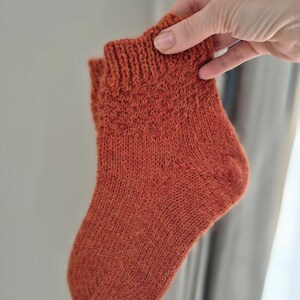May include: A hand-knitted, rust-colored sock. The sock is held up, showcasing its textured knit pattern and ribbed cuff. The warm, earthy tone suggests a cozy, handmade item, perfect for cold weather.