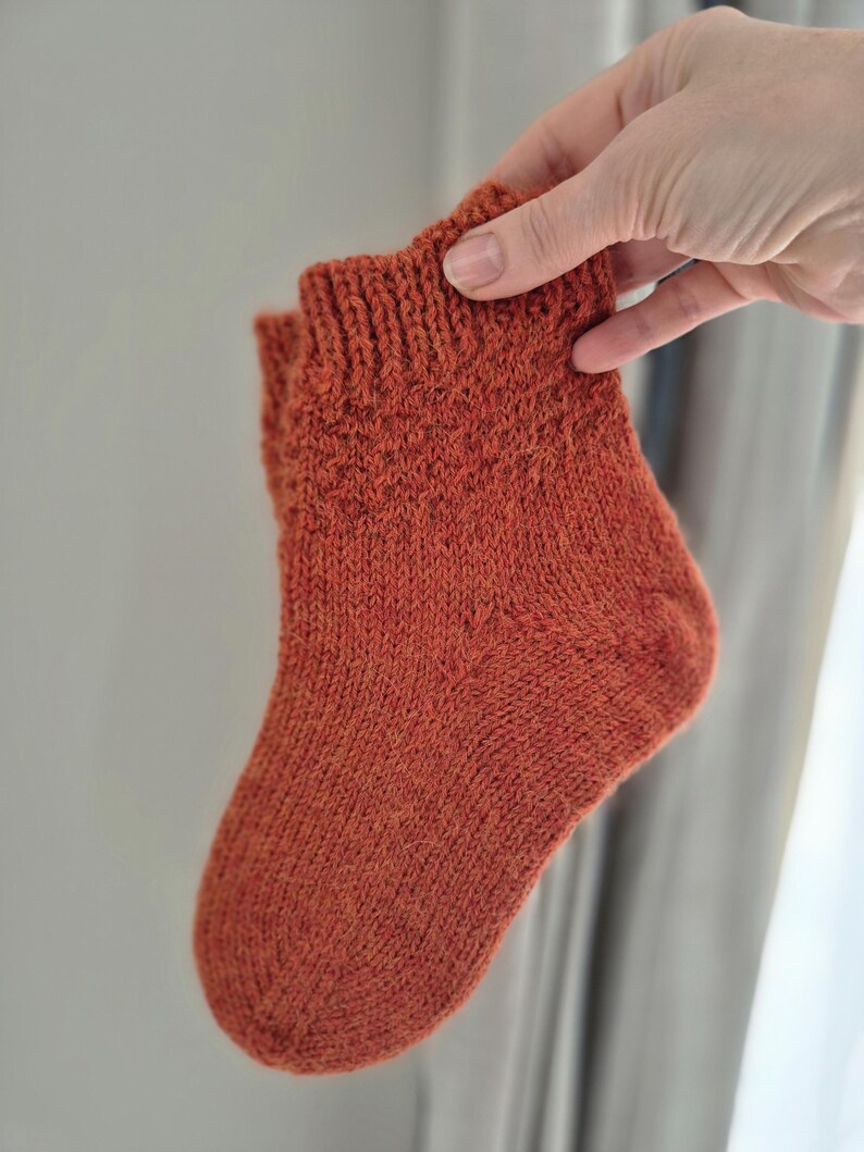 May include: A hand-knitted, rust-colored sock. The sock is held up, showcasing its textured knit pattern and ribbed cuff. The warm, earthy tone suggests a cozy, handmade item, perfect for cold weather.