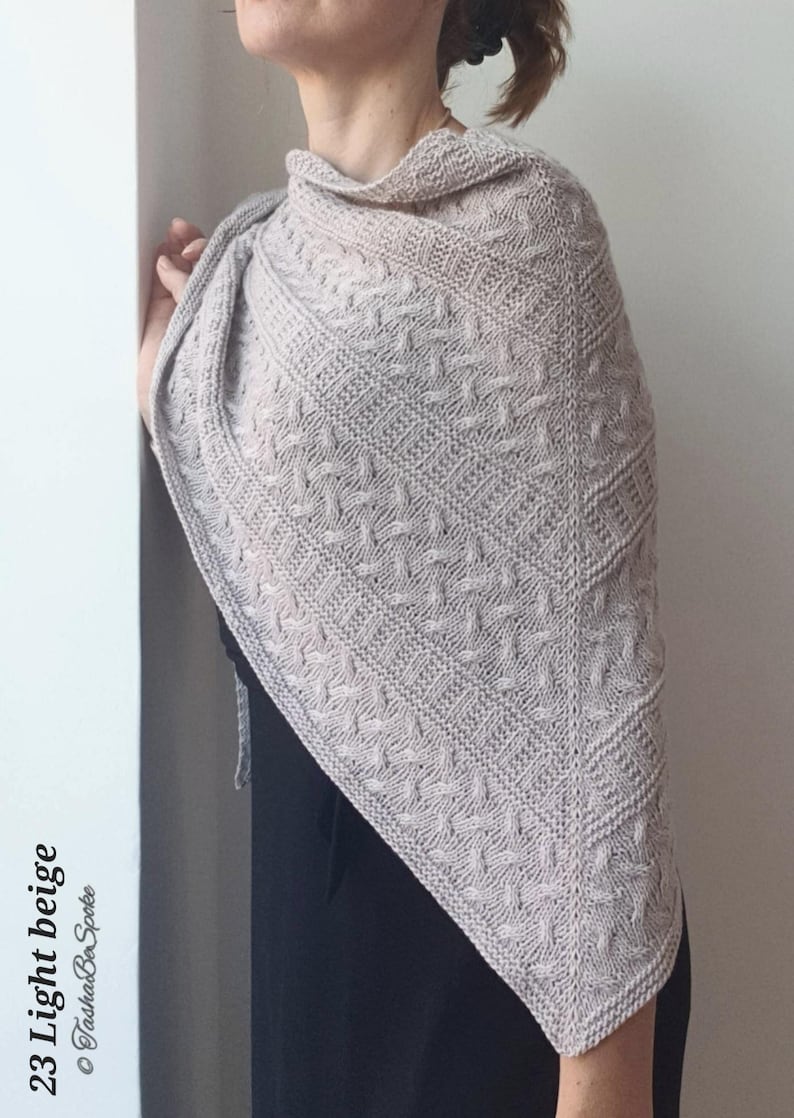 May include: A light beige knitted shawl with a cable stitch pattern. The shawl is draped over a person's shoulders.