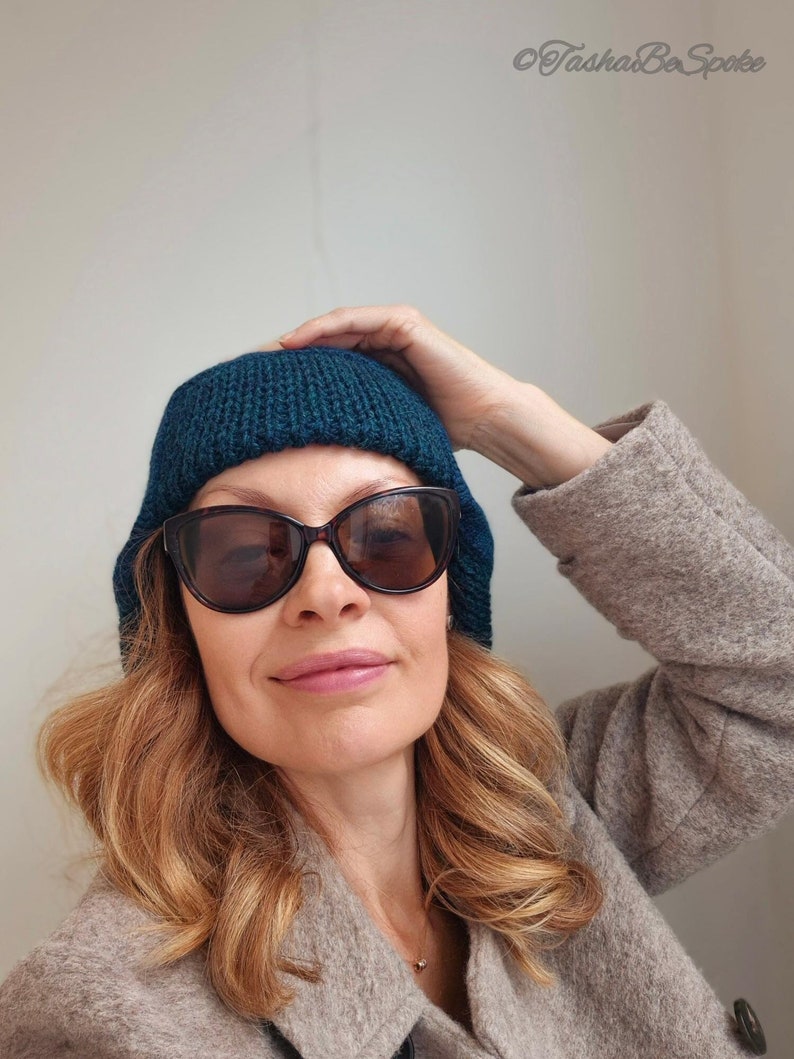 K&ouml;nnte beinhalten: Eine Frau tr&auml;gt eine tealblaue Strickm&uuml;tze und eine braune Sonnenbrille. Sie tr&auml;gt einen hellbraunen Mantel.