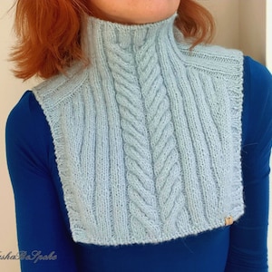 May include: A light blue knitted cowl with a cable knit pattern. The cowl is worn over a blue shirt.