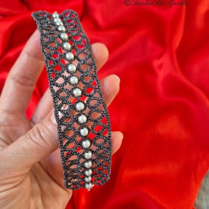 Puede incluir: Un brazalete de encaje negro con perlas blancas. El brazalete est&aacute; hecho de encaje de ganchillo delicado y presenta una fila de perlas en el centro.
