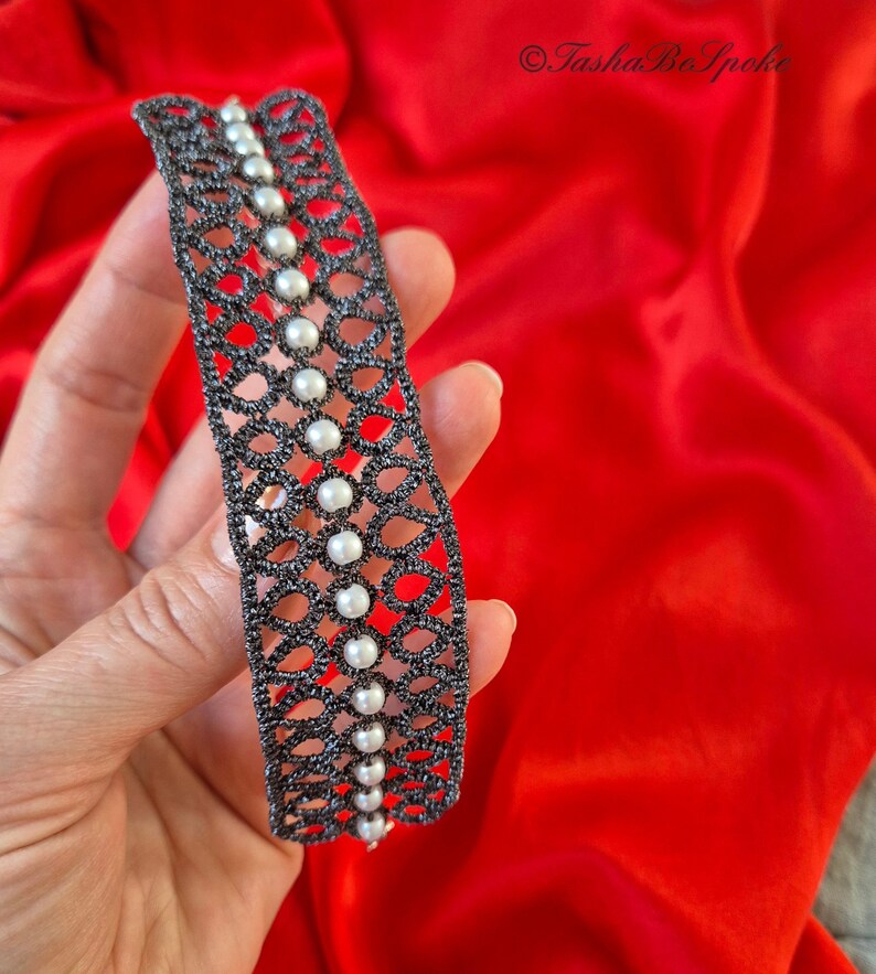 Puede incluir: Un brazalete de encaje negro con perlas blancas. El brazalete est&aacute; hecho de encaje de ganchillo delicado y presenta una fila de perlas en el centro.