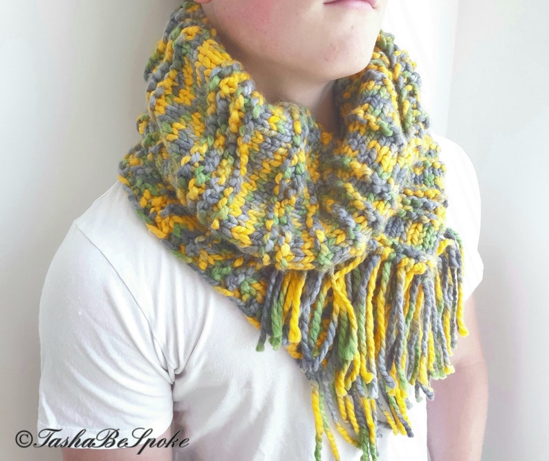 May include: A gray, yellow, and green knitted cowl with long fringe. The cowl is worn by a person.