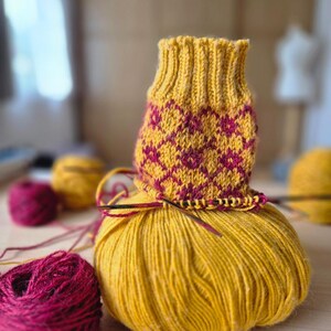 May include: A partially knitted yellow and burgundy sock sits atop a large ball of yellow yarn. The sock features a geometric pattern in burgundy. Additional burgundy yarn and a yellow yarn ball are in the background, along with knitting needles.