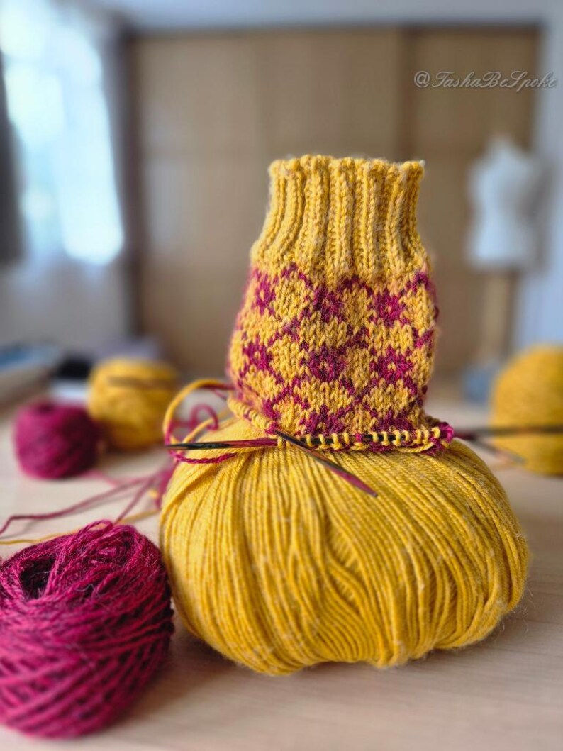 May include: A partially knitted yellow and burgundy sock sits atop a large ball of yellow yarn. The sock features a geometric pattern in burgundy. Additional burgundy yarn and a yellow yarn ball are in the background, along with knitting needles.