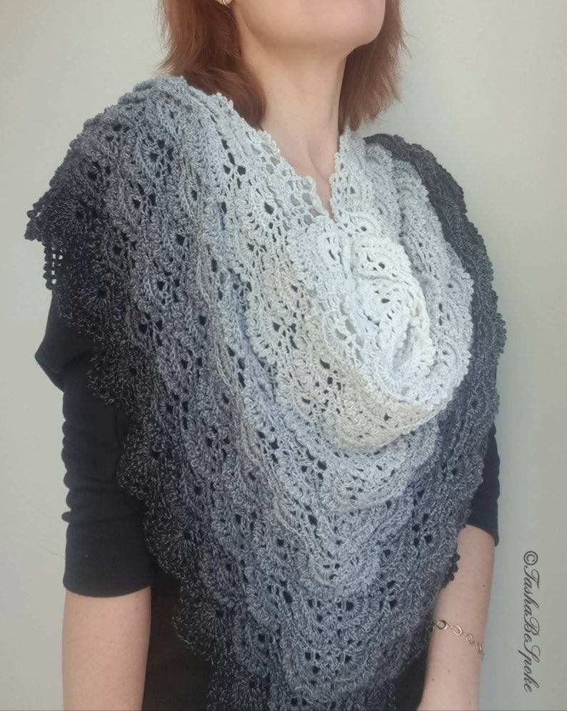 May include: A grey and white crochet shawl with a delicate lace pattern. The shawl is draped around a person's shoulders and neck.