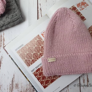 K&ouml;nnte beinhalten: Eine rosafarbene Strickm&uuml;tze mit einem kleinen goldenen Etikett mit der Aufschrift "Fasha Be Spoke". Die M&uuml;tze liegt auf einem Buch mit einem rot-wei&szlig;en Muster.