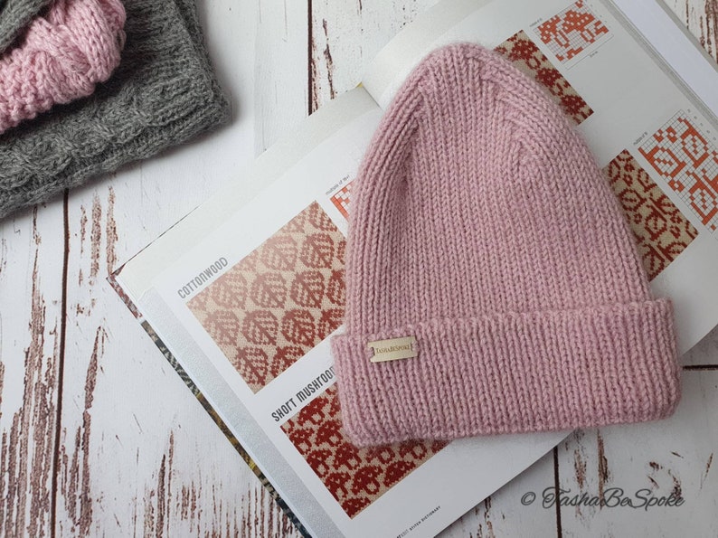 K&ouml;nnte beinhalten: Eine rosafarbene Strickm&uuml;tze mit einem kleinen goldenen Etikett mit der Aufschrift "Fasha Be Spoke". Die M&uuml;tze liegt auf einem Buch mit einem rot-wei&szlig;en Muster.