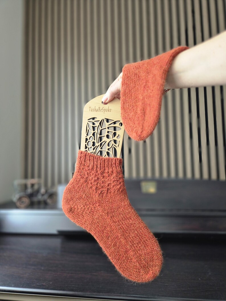 May include: A pair of orange knitted socks, one on a wooden display with the text "TashaBeSpoke". The socks are a warm, earthy tone, with a textured knit pattern. The display has a floral cut-out design.