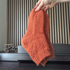 May include: A pair of orange knitted socks. The socks are held up by a hand, showcasing their textured knit design and warm color. The socks appear to be handmade, with a focus on comfort and style.