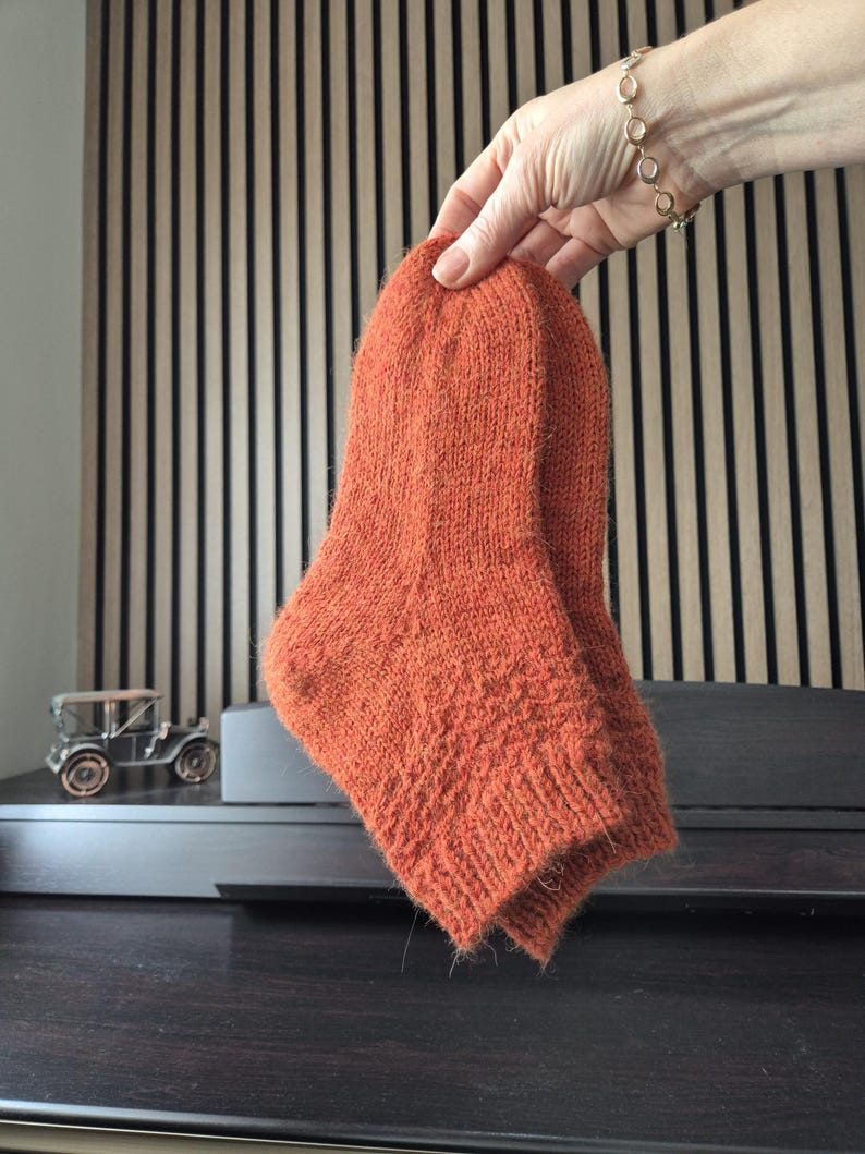 May include: A pair of orange knitted socks. The socks are held up by a hand, showcasing their textured knit design and warm color. The socks appear to be handmade, with a focus on comfort and style.