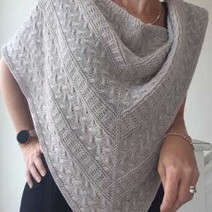 May include: A gray knitted shawl with a cable knit pattern. The shawl is draped over a person's shoulders and is worn with a black top.