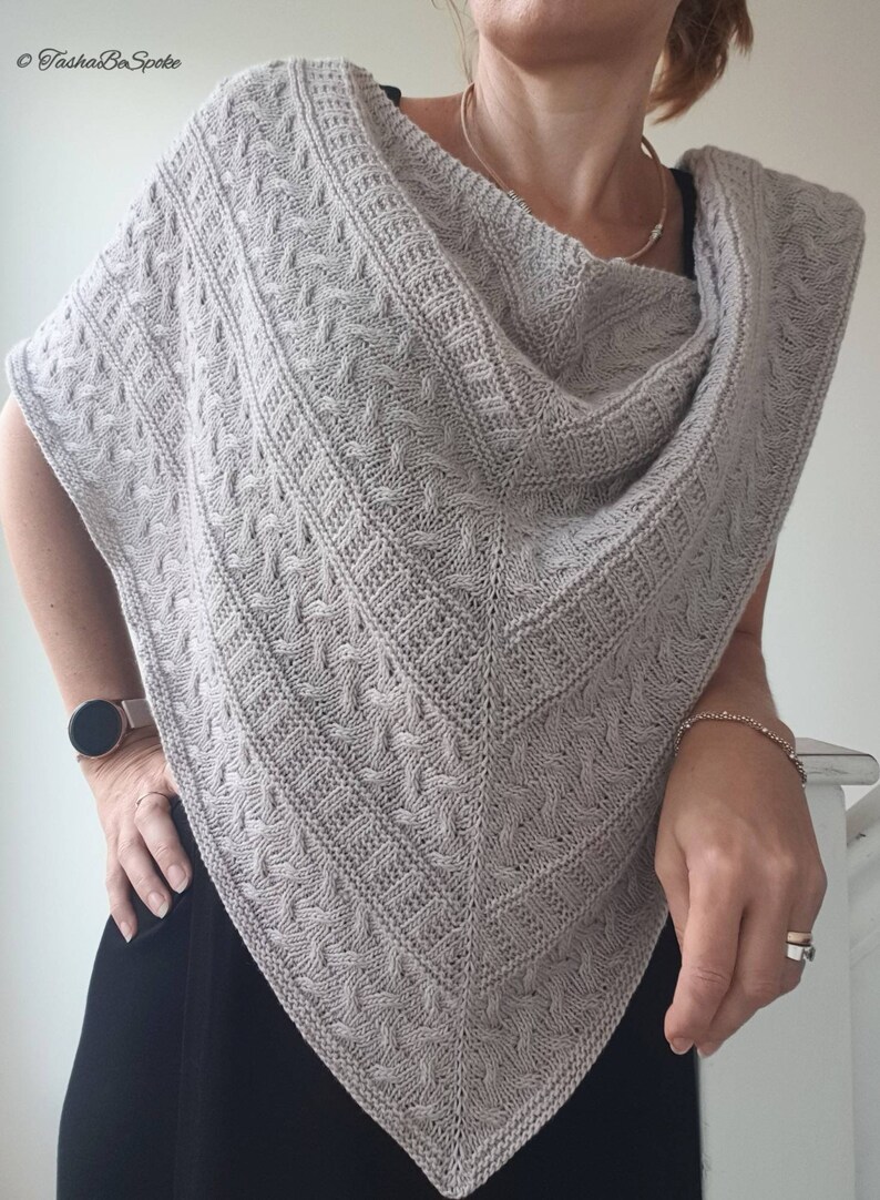 May include: A gray knitted shawl with a cable knit pattern. The shawl is draped over a person's shoulders and is worn with a black top.