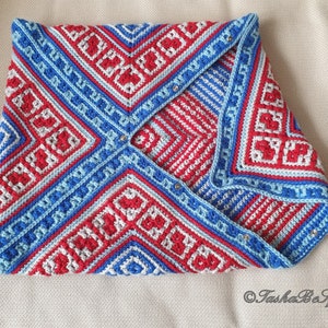 Op de afbeelding: Een gevouwen, gehaakt item met een geometrisch patroon in rood, wit en blauw. Het ontwerp heeft vierkanten en lijnen, met een lichtblauwe rand. Het item is vastgezet met kleine, zilverkleurige bevestigingsmiddelen. Het algemene thema is een handgemaakt textiel.