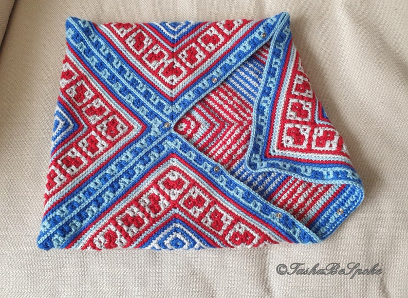 Op de afbeelding: Een gevouwen, gehaakt item met een geometrisch patroon in rood, wit en blauw. Het ontwerp heeft vierkanten en lijnen, met een lichtblauwe rand. Het item is vastgezet met kleine, zilverkleurige bevestigingsmiddelen. Het algemene thema is een handgemaakt textiel.