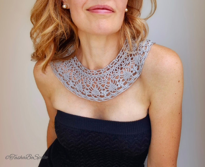 May include: A gray crocheted collar necklace with a delicate floral pattern. The necklace is worn by a person with light brown hair and a black strapless top.