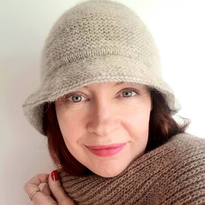 May include: A light beige knitted cloche hat with a small brim. The hat is made of a soft, textured yarn. A matching brown scarf is wrapped around the neck. The woman is wearing a red top.
