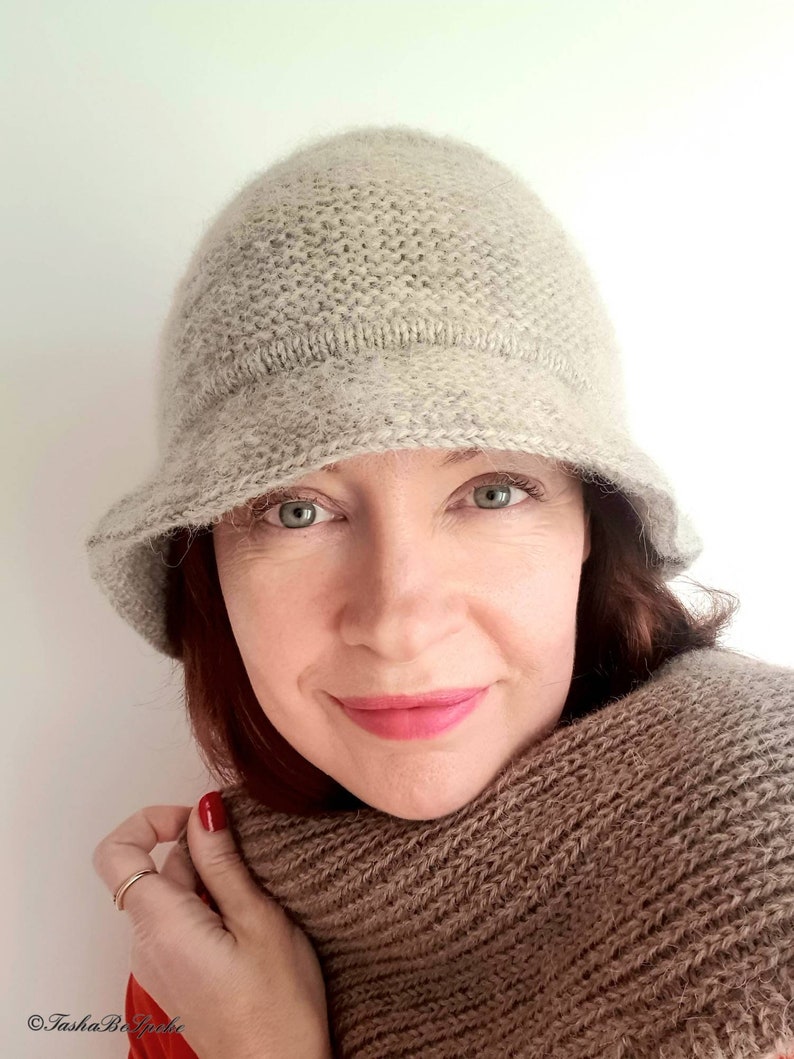 May include: A light beige knitted cloche hat with a small brim. The hat is made of a soft, textured yarn. A matching brown scarf is wrapped around the neck. The woman is wearing a red top.