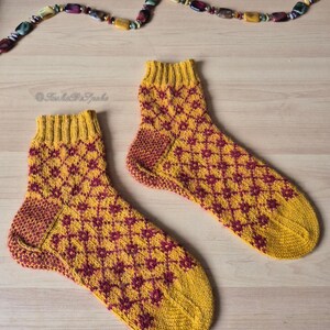 May include: A pair of hand-knitted socks with a yellow and burgundy geometric pattern. The socks have a ribbed cuff and heel, and a patterned design on the foot. The socks are displayed on a light wooden surface.