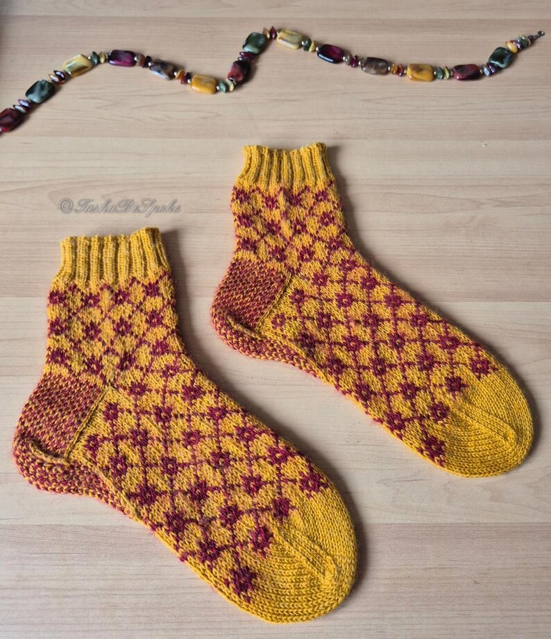 May include: A pair of hand-knitted socks with a yellow and burgundy geometric pattern. The socks have a ribbed cuff and heel, and a patterned design on the foot. The socks are displayed on a light wooden surface.
