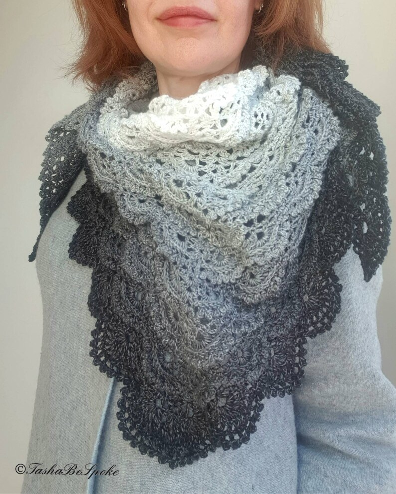 May include: A black and white ombre crochet shawl with a delicate lace pattern. The shawl is draped around a person's neck and shoulders.