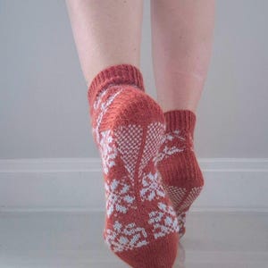 May include: A pair of knitted socks in a rust-colored hue, adorned with a white floral pattern. The socks are visible from the ankles down, showcasing the intricate design and texture of the knitwear. The socks are on a white surface.