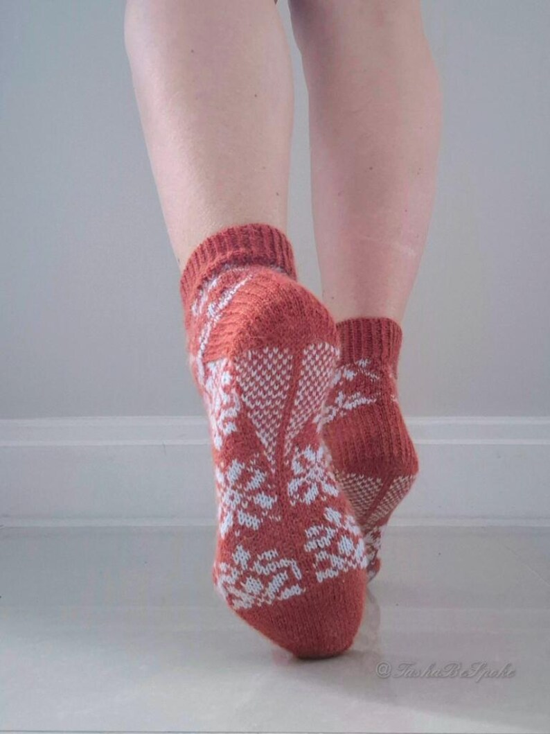 May include: A pair of knitted socks in a rust-colored hue, adorned with a white floral pattern. The socks are visible from the ankles down, showcasing the intricate design and texture of the knitwear. The socks are on a white surface.
