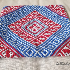 Op de afbeelding: Een handgemaakte gehaakte kussensloop met een geometrisch ontwerp in rood, wit en blauw. Het patroon bevat ruit- en vierkantvormen, waardoor een textuur ontstaat. De kussensloop is vierkant van vorm.
