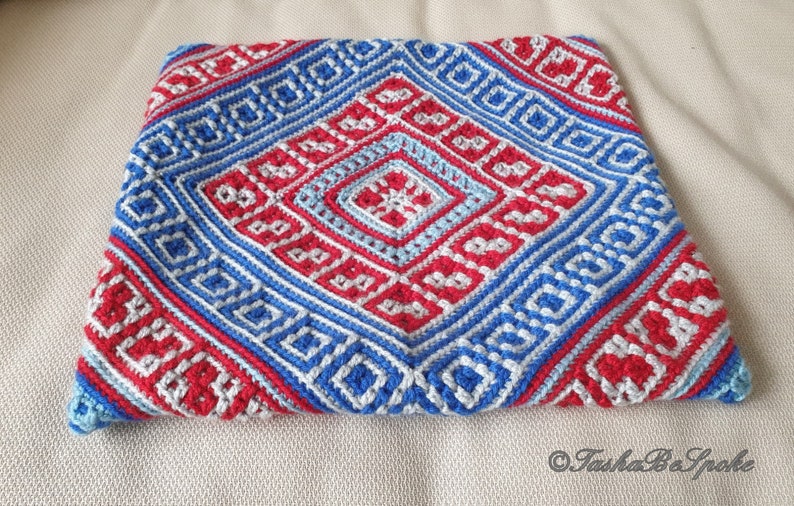 Op de afbeelding: Een handgemaakte gehaakte kussensloop met een geometrisch ontwerp in rood, wit en blauw. Het patroon bevat ruit- en vierkantvormen, waardoor een textuur ontstaat. De kussensloop is vierkant van vorm.