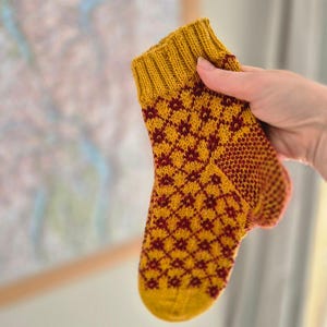 May include: A hand-knitted sock in shades of gold and burgundy. The sock features a diamond pattern and ribbed cuff. The sock is held up against a blurred background. The sock is a small size, likely for a baby or child.