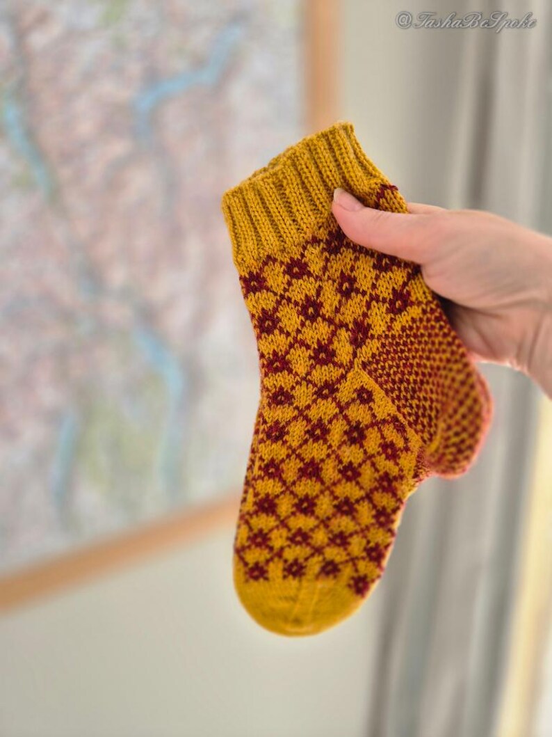 May include: A hand-knitted sock in shades of gold and burgundy. The sock features a diamond pattern and ribbed cuff. The sock is held up against a blurred background. The sock is a small size, likely for a baby or child.