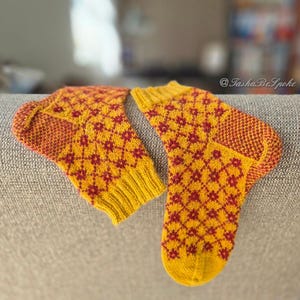 May include: A pair of hand-knitted socks in yellow and red. The socks feature a repeating diamond pattern in red against a yellow background. The heels and toes are a darker shade of red. The socks are resting on a gray surface.