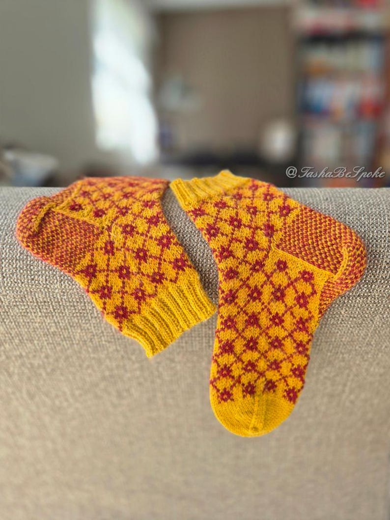 May include: A pair of hand-knitted socks in yellow and red. The socks feature a repeating diamond pattern in red against a yellow background. The heels and toes are a darker shade of red. The socks are resting on a gray surface.