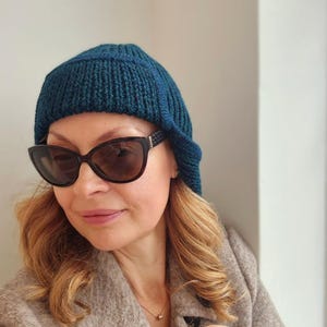 K&ouml;nnte beinhalten: Eine Frau tr&auml;gt eine tealblaue Strickm&uuml;tze mit einem langen Lappen im R&uuml;cken, eine schwarze Sonnenbrille und einen braunen Mantel.