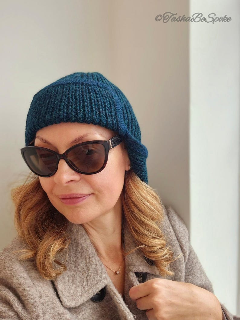 K&ouml;nnte beinhalten: Eine Frau tr&auml;gt eine tealblaue Strickm&uuml;tze mit einem langen Lappen im R&uuml;cken, eine schwarze Sonnenbrille und einen braunen Mantel.