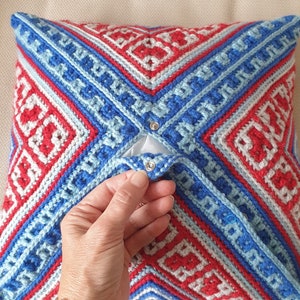 Op de afbeelding: Een vierkant, gehaakt kussen met een geometrisch patroon in rood, wit en blauw. Het kussen heeft een flap die met zilveren drukknopen is vastgezet. Het ontwerp heeft ruit- en rechthoekige vormen. Een decoratief woonaccessoire.