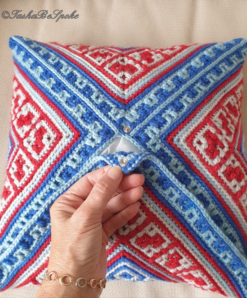 Op de afbeelding: Een vierkant, gehaakt kussen met een geometrisch patroon in rood, wit en blauw. Het kussen heeft een flap die met zilveren drukknopen is vastgezet. Het ontwerp heeft ruit- en rechthoekige vormen. Een decoratief woonaccessoire.