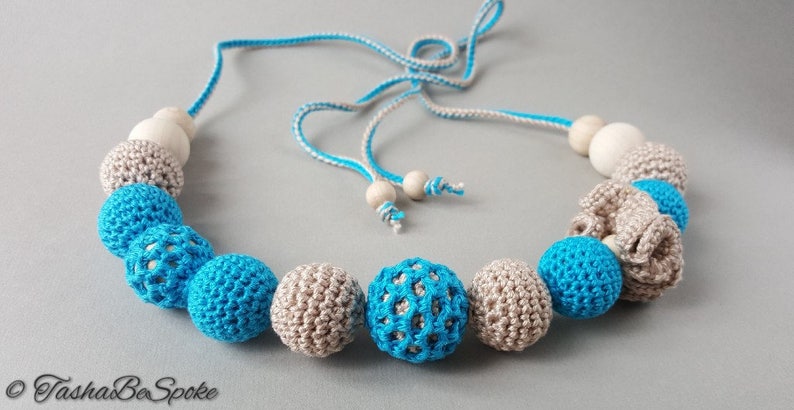 Op de afbeelding: Een handgemaakte ketting met gehaakte kralen in blauwe en beige tinten, afgewisseld met houten kralen. De ketting heeft een blauw-wit koord en is verstelbaar. Het ontwerp omvat open en effen gehaakte bollen.
