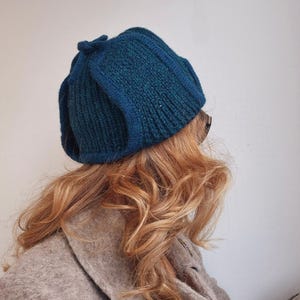 K&ouml;nnte beinhalten: Eine tealblaue Strickm&uuml;tze mit einem einzigartigen gefalteten Design. Die M&uuml;tze ist aus einem weichen, strukturierten Garn gefertigt.