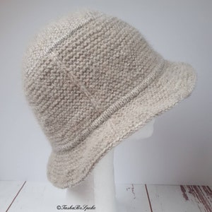 May include: A light beige knitted cloche hat with a small brim. The hat is displayed on a white mannequin head against a white and wood-grain background. The hat has a textured appearance and a rounded crown.