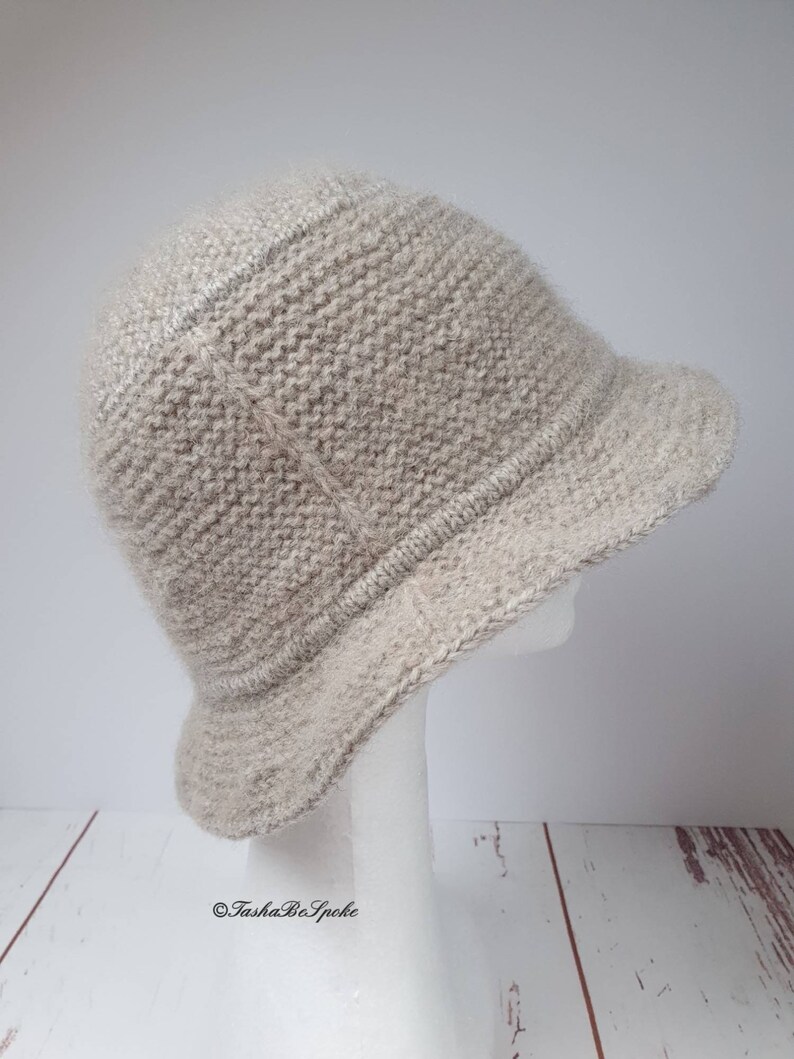 May include: A light beige knitted cloche hat with a small brim. The hat is displayed on a white mannequin head against a white and wood-grain background. The hat has a textured appearance and a rounded crown.