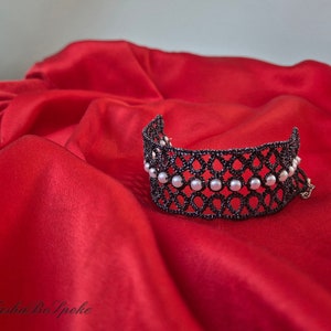 Puede incluir: Un brazalete de encaje negro con detalles de perlas blancas sobre un fondo de sat&eacute;n rojo.