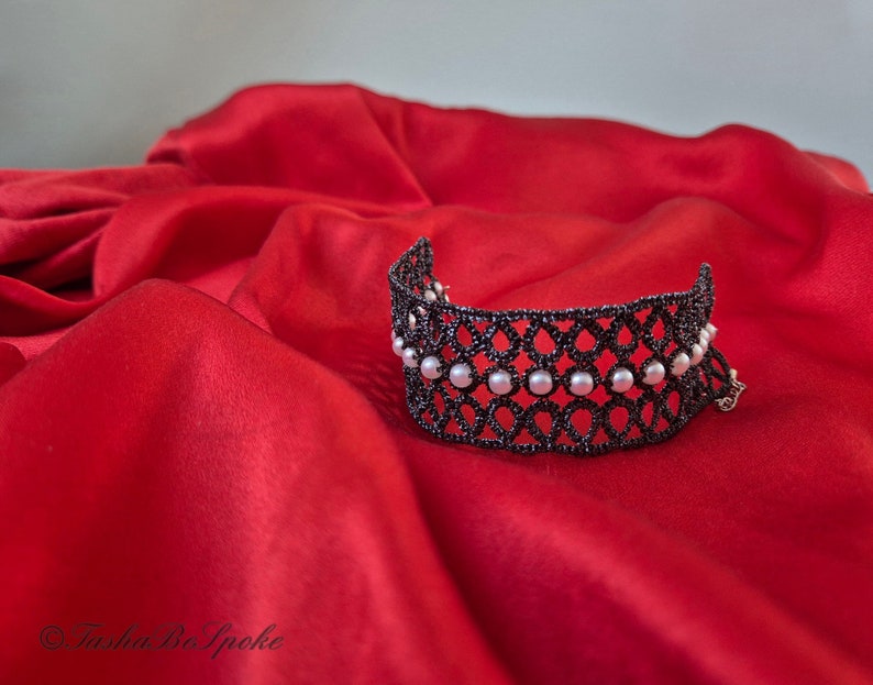 Puede incluir: Un brazalete de encaje negro con detalles de perlas blancas sobre un fondo de sat&eacute;n rojo.