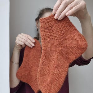 May include: A pair of hand-knitted socks in a warm orange hue. The socks are held up, showcasing their textured knit pattern and detailed cuff design. The yarn appears soft and cozy, perfect for cold weather.