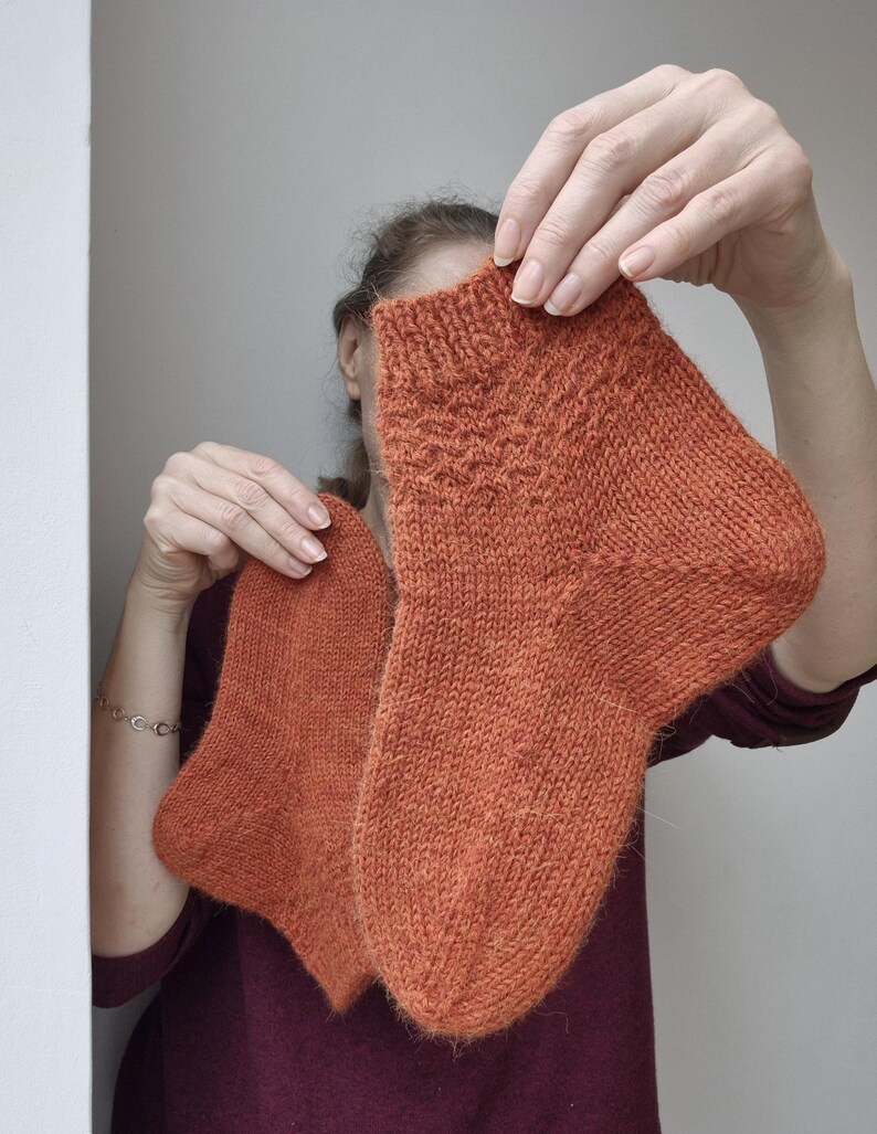 May include: A pair of hand-knitted socks in a warm orange hue. The socks are held up, showcasing their textured knit pattern and detailed cuff design. The yarn appears soft and cozy, perfect for cold weather.
